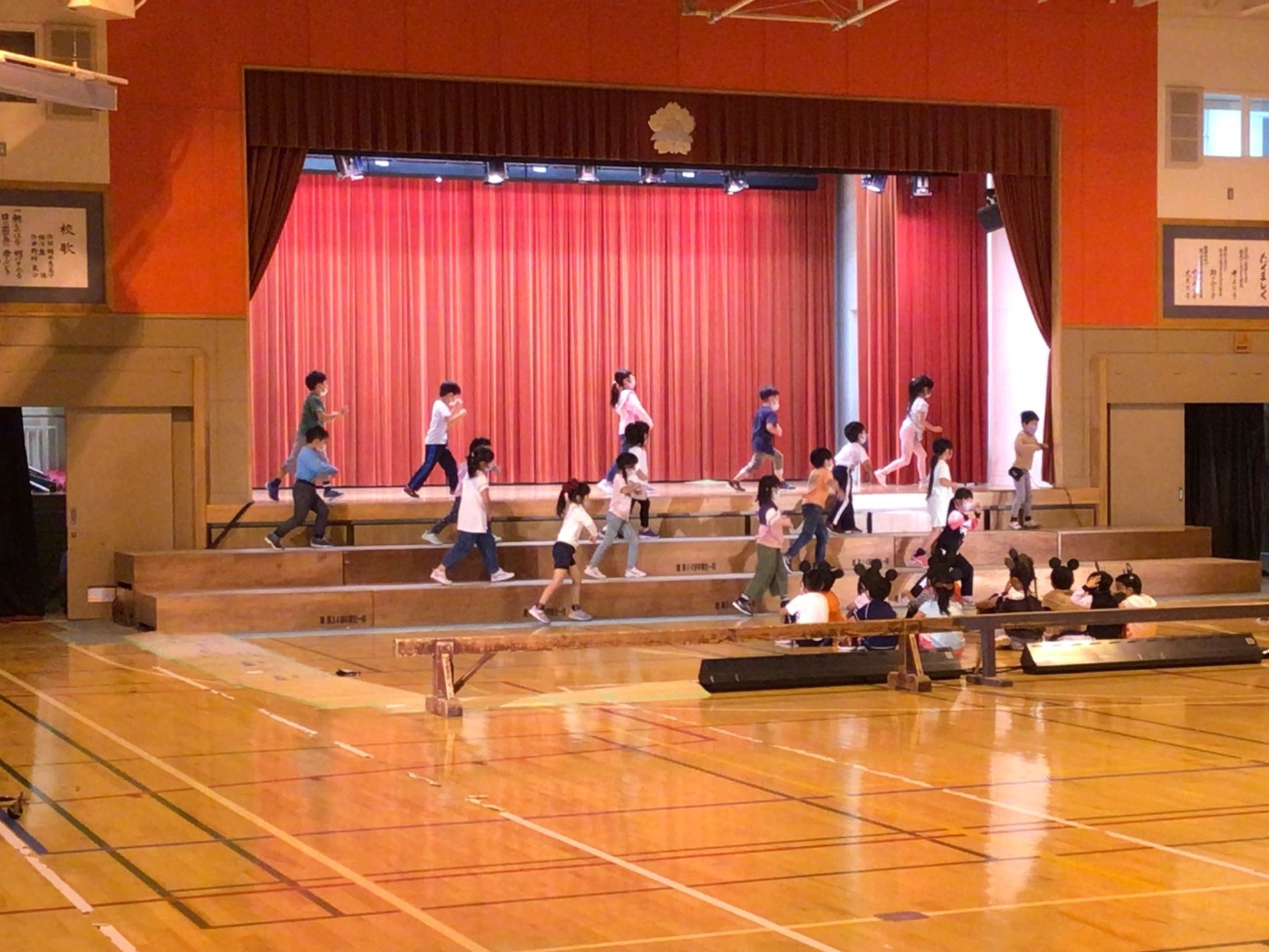 体育館の舞台で四列になり手を前に出して踊りの練習をしている子供たちと、それを間近で見ている体育座りをした子供たちの写真
