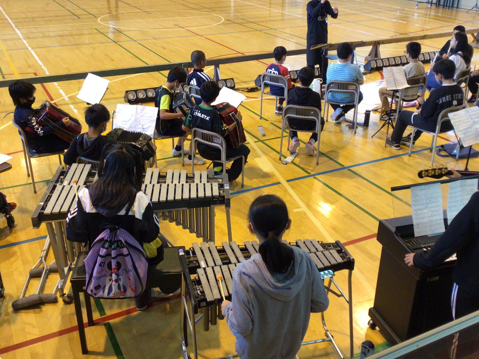 体育館に並べられた椅子に座り、指揮者に合わせて様々な楽器を練習する子供たちの写真