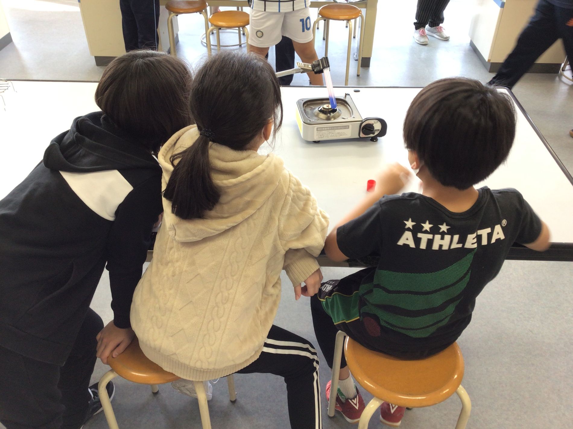 ガスコンロに試験管を置いて加熱している様子を机に座って眺めている3人の子どもたちの写真