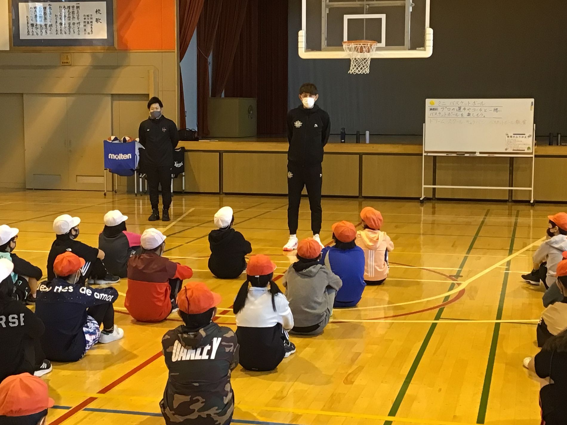 体育館に黒いジャージ姿の男性が立ち、向かい合うようにして、白やオレンジの帽子をかぶった子どもたちが床に座っている様子の写真