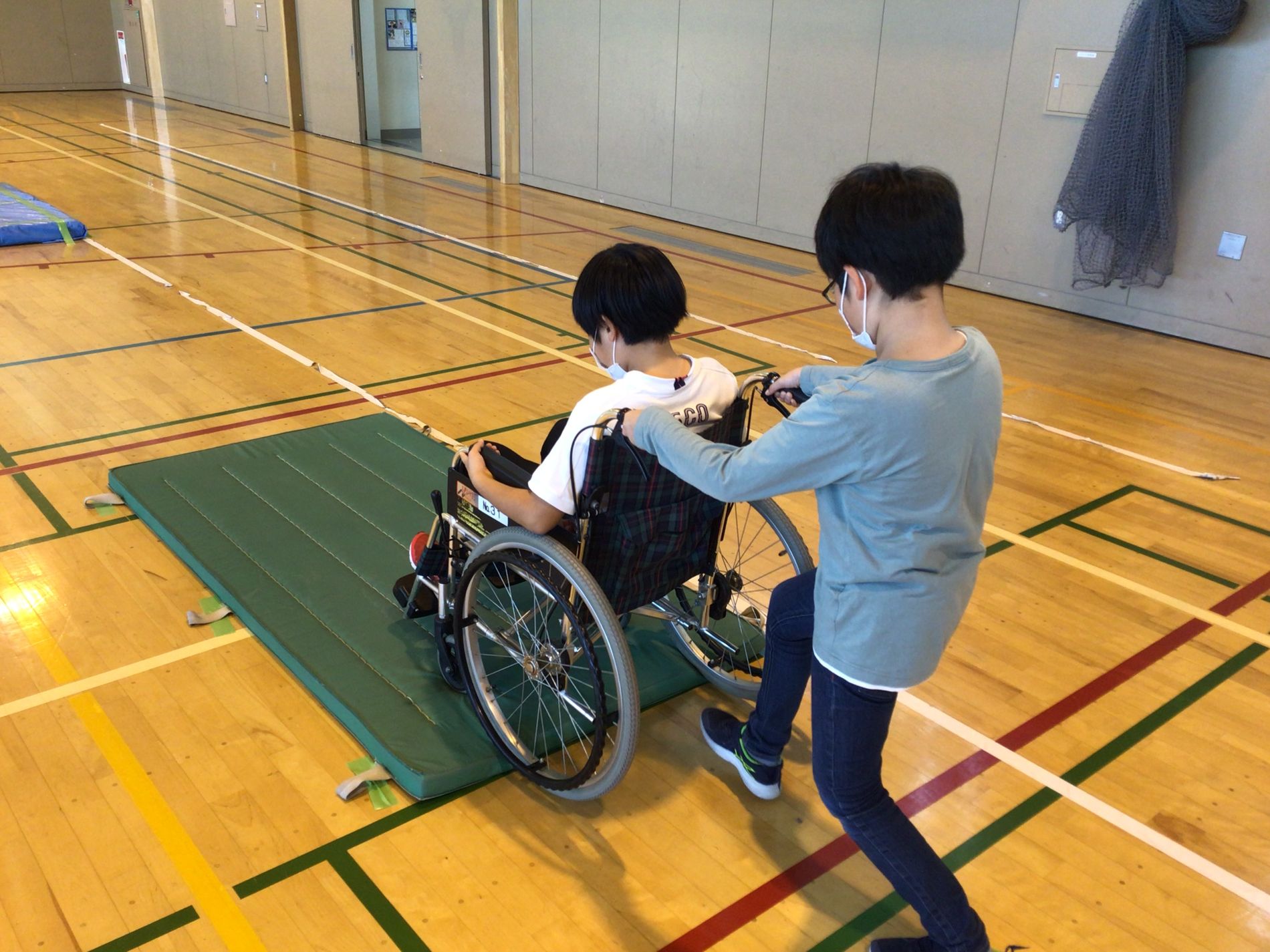 緑色のマットの上で、車椅子に座った男の子を膝を曲げながら押し、段差を乗り越える練習をしている男の子の写真