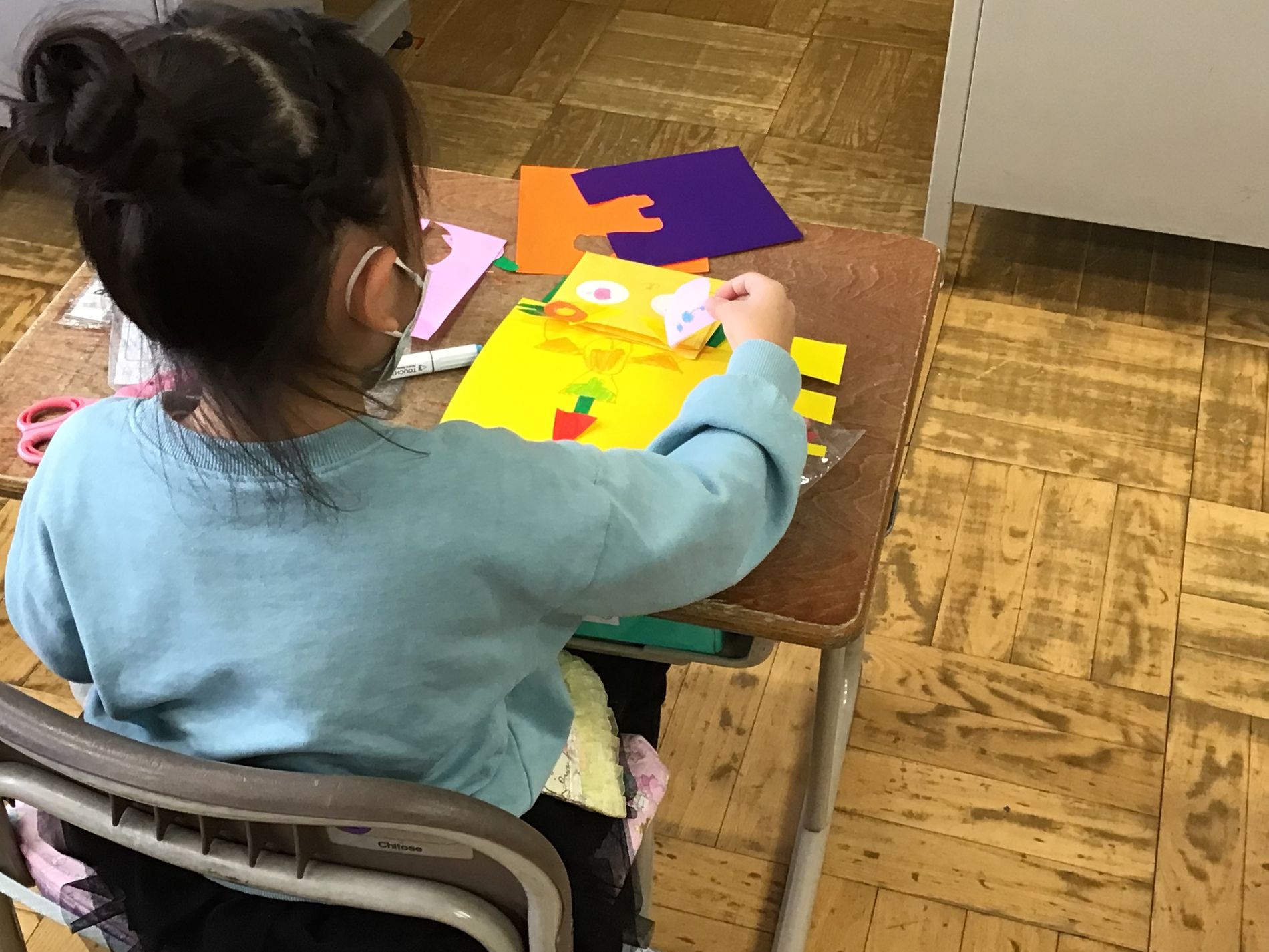 机に座って黄色い折り紙に目や耳などのパーツを配置してみている女の子の写真
