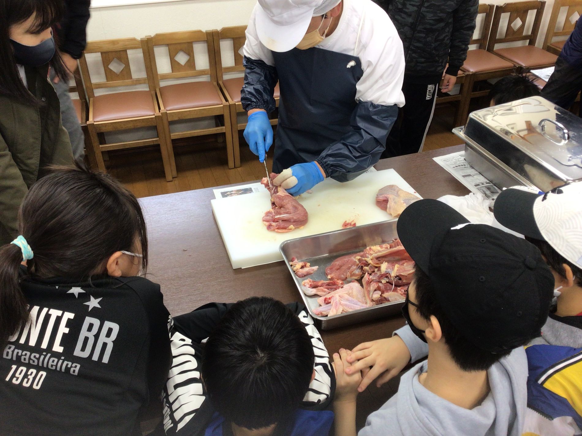 エプロンを付けた一人の男性が、包丁とまな板で生肉をさばいている様子を興味津々に見つめている子どもたちの写真