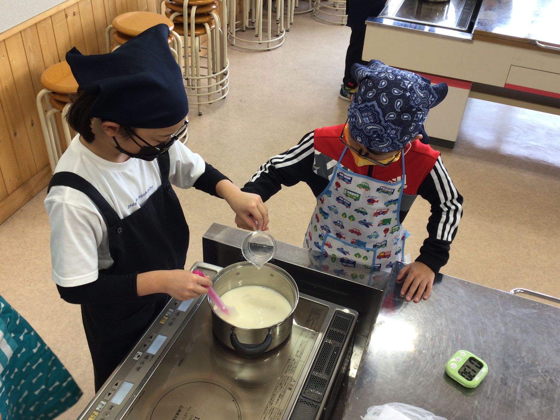 IHコンロに豆腐の素が入った鍋を火にかけながら、計量カップに入ったにがりを一緒に鍋に入れているエプロン姿の2人の生徒の写真