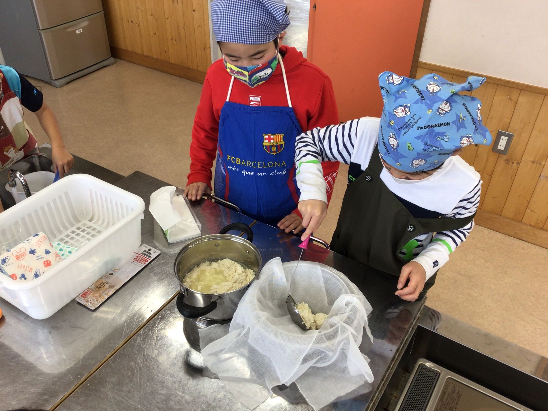 鍋にできた豆腐を、お玉を使ってザルに濾している男の子とそれを見守る男の子の写真